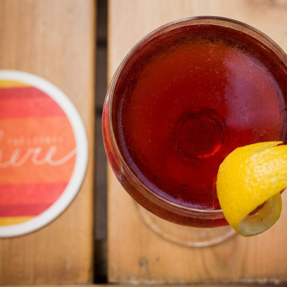 Top-down view of a red cocktail garnished with a lemon peel on a wooden table beside a colorful circular coaster.
