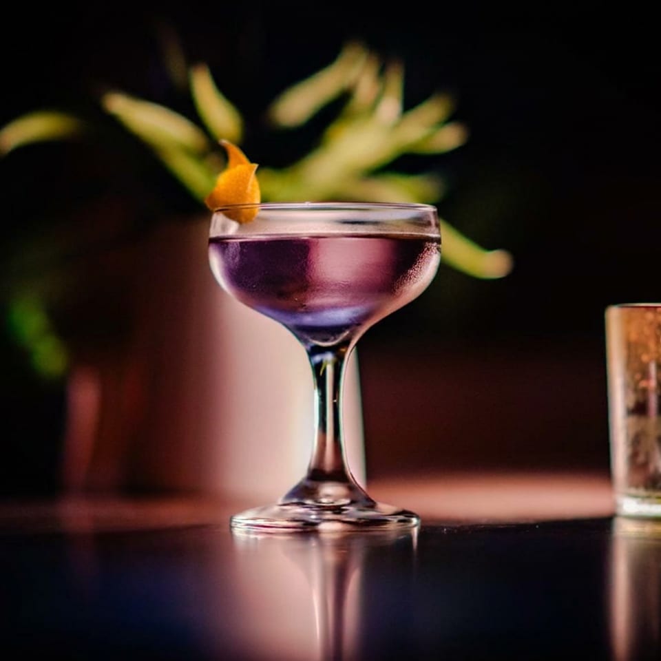 Purple cocktail in a coupe glass garnished with an orange twist, set on a reflective bar in a dimly lit setting.