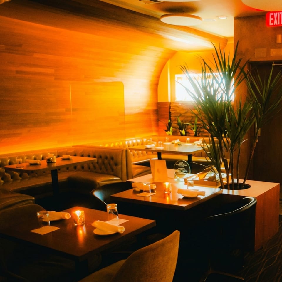 Warmly lit restaurant dining room with leather booths, set tables, candles, tall plants, and wood-paneled walls.