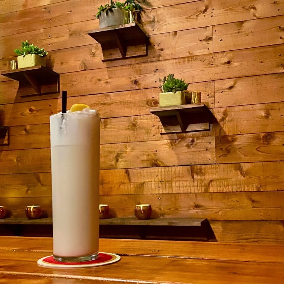 Tall creamy cocktail garnished with an orange slice on a coaster, set against a warm wooden wall with plants and candles.