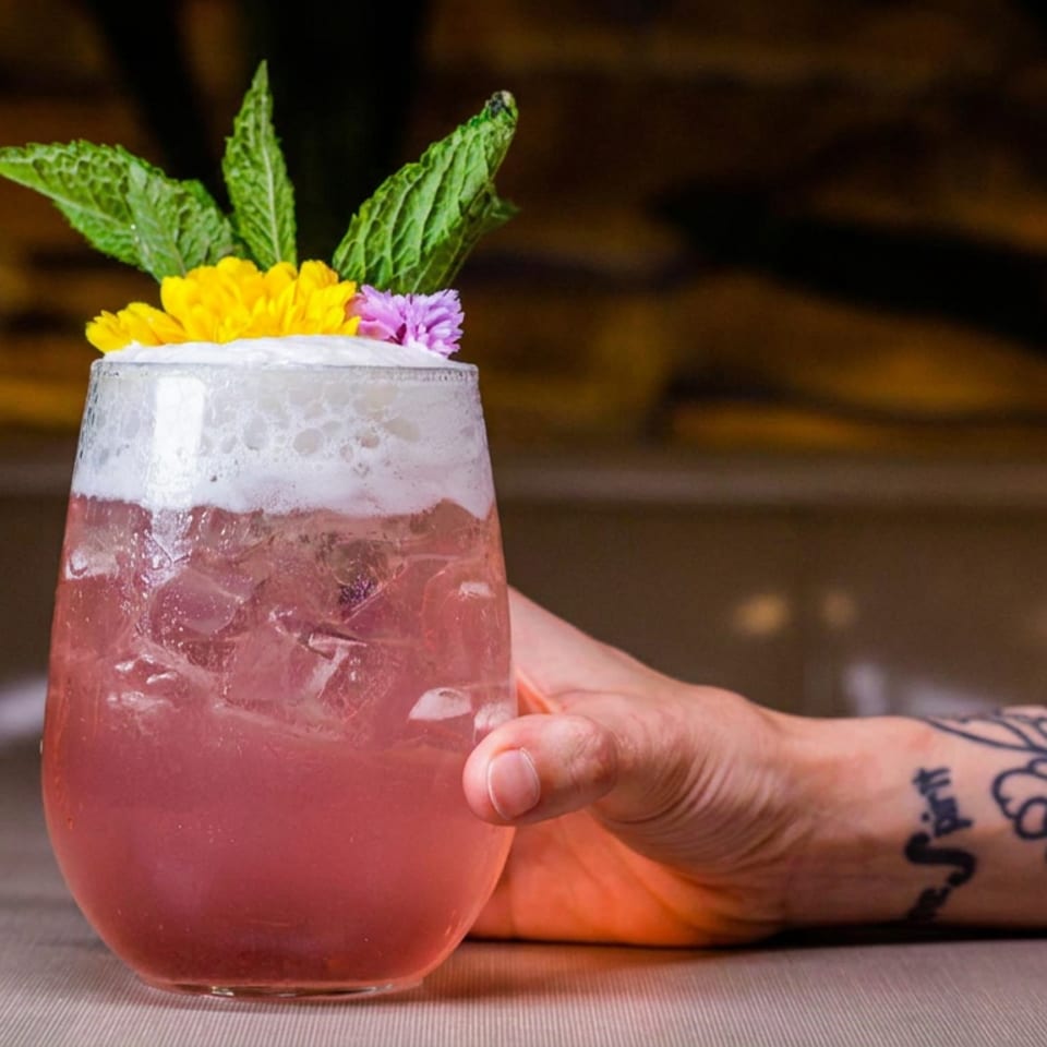 Pink iced cocktail topped with foam, mint leaves, and colorful flowers, held by a tattooed arm at a restaurant bar.
